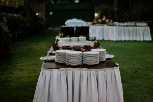 Een stapel borden staat op een tafel met een wit tafelkleed. De tafel staat buiten op het gras en het is avond.