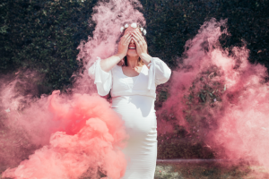 Een zwangere vrouw in een witte jurk houdt haar handen voor haar ogen terwijl om haar heen roze kleurstof in de lucht wordt gegooid.