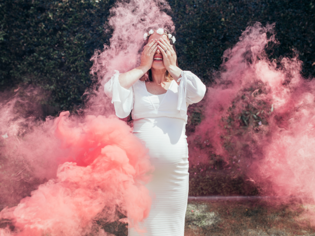 Een zwangere vrouw in een witte jurk houdt haar handen voor haar ogen terwijl om haar heen roze kleurstof in de lucht wordt gegooid.