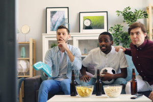 Drie mannen zitten op een bank en kijken gespannen naar een tv-scherm. Er staan drie bierflessen en twee kommen met popcorn en chips op de tafel voor hen.