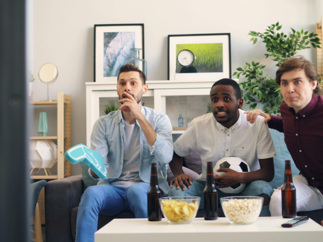 Drie mannen zitten op een bank en kijken gespannen naar een tv-scherm. Er staan drie bierflessen en twee kommen met popcorn en chips op de tafel voor hen.
